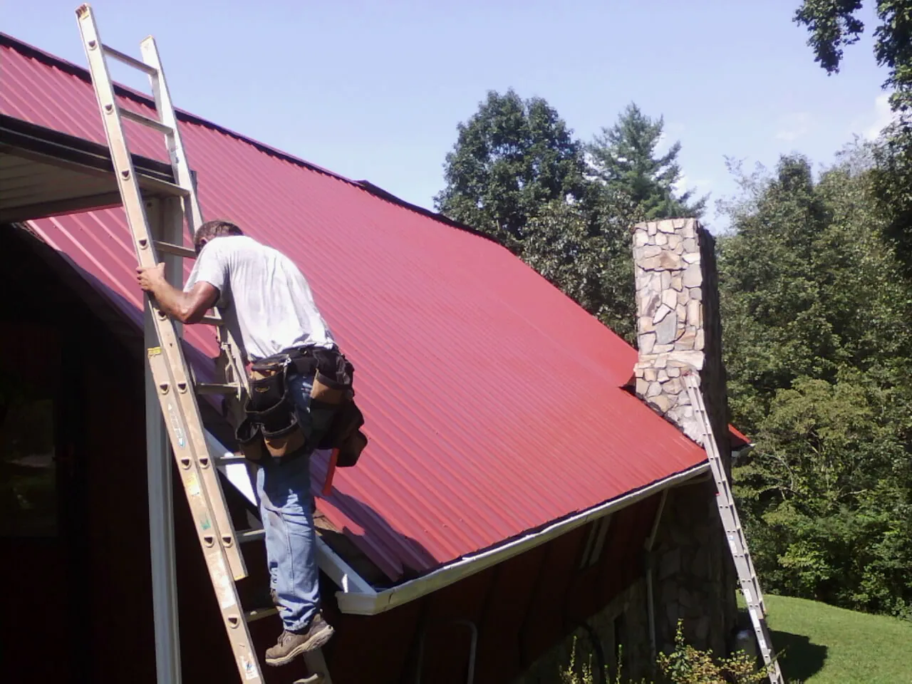 Roofer on a ladder beside a red metal roof and stone chimney — Soffit & Fascia Repair by Roofing Brothers in Pierceton